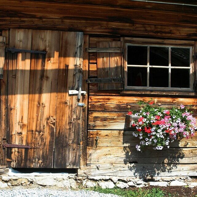 Eine alte Holzhütte von der Vorderansicht, den Eingangsbereich und Blumen im Trog laden ein mehr dort zu entdecken.